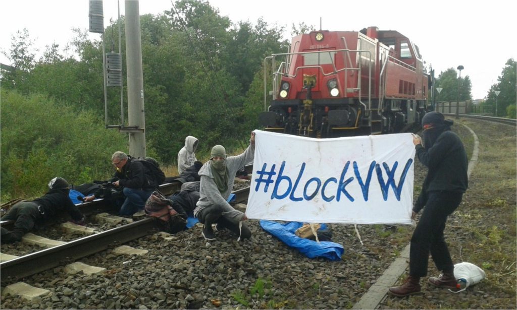Aktivist*innen, die vor einem blockierten Zug angekettet sind. Auf ihrem Transpi steht #blockVW.